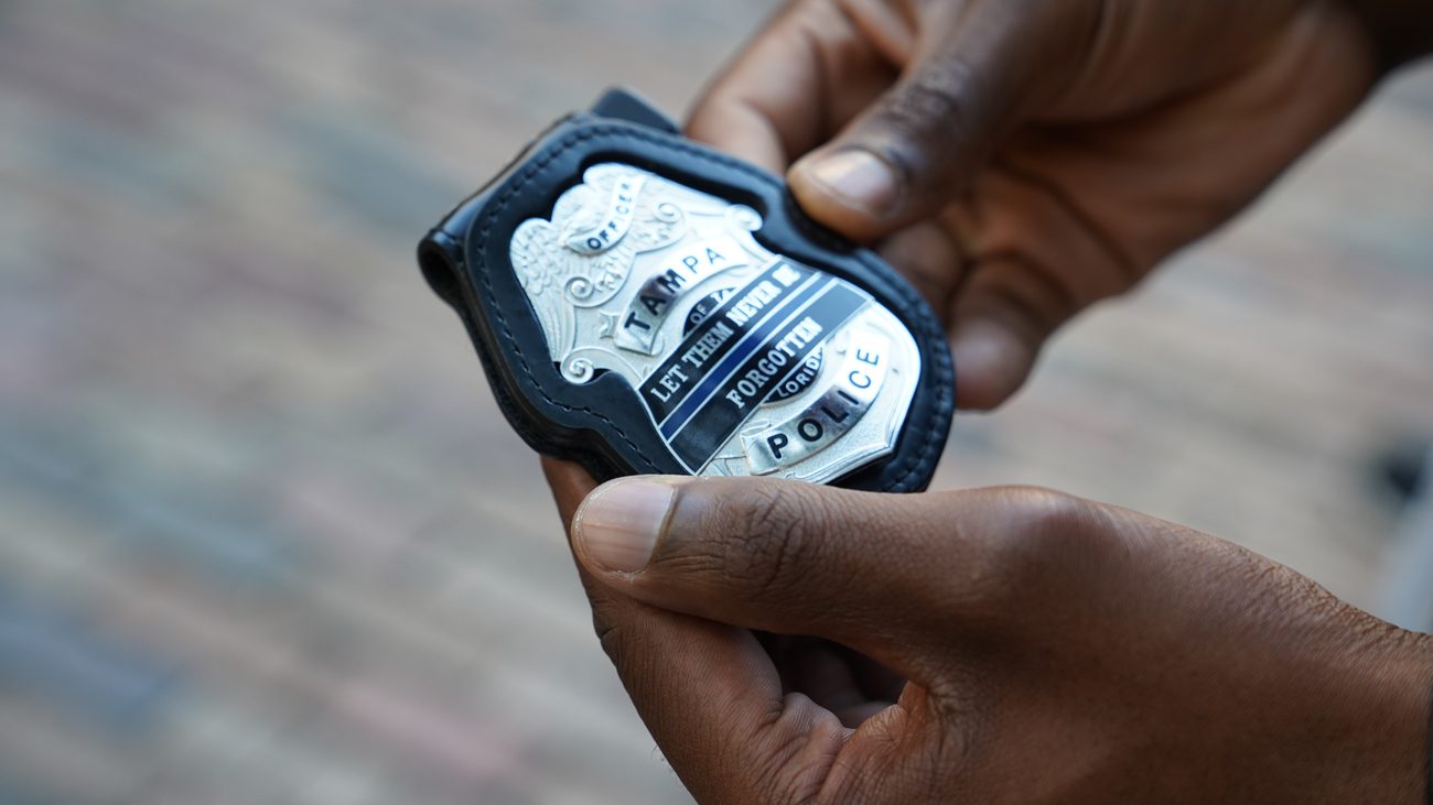 Hands holding the Tampa Police memorial badge inscribed Let Them Never Be Forgotten