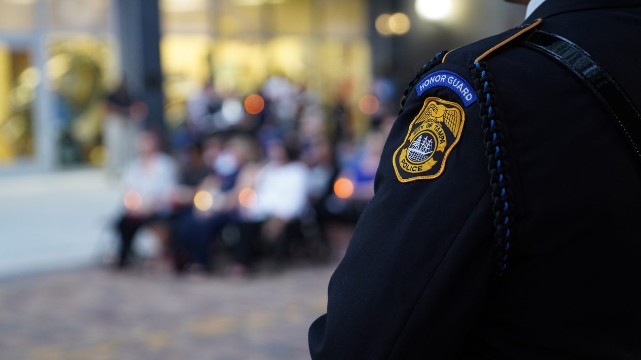 A Tampa Police honor guard member at a memorial ceremony, with those gathered seated behind